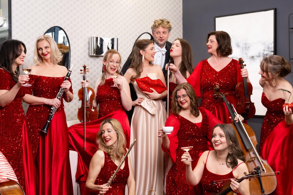Eine Gruppe von Frauen in roten Abendkleidern steht zusammen und posiert fröhlich. Ein Mann ist ebenfalls dabei, während einige Frauen Instrumente halten und Cocktails trinken. | © Michaela Begsteiger