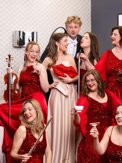 Eine Gruppe von Frauen in roten Abendkleidern steht zusammen und posiert fröhlich. Ein Mann ist ebenfalls dabei, während einige Frauen Instrumente halten und Cocktails trinken. | © Michaela Begsteiger