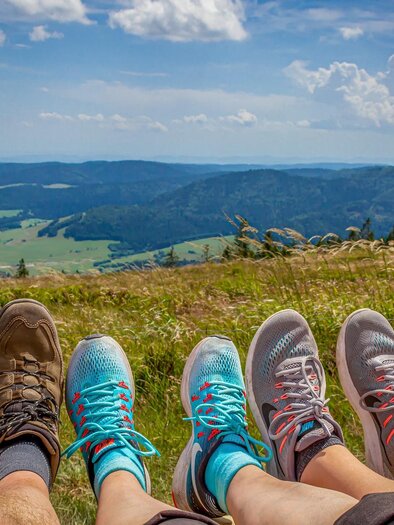 Vier Paare von Füßen stehen auf einer grünen Wiese mit einer Aussicht auf Berge und einen blauen Himmel. Die Szene strahlt Entspannung und Naturverbundenheit aus. | © pixabay.com