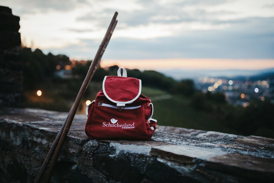 Ein roter Rucksack steht auf einer Steinmauer, neben einem Wanderstock. Im Hintergrund ist ein schöner Abendhimmel zu sehen, der die Stadtbeleuchtung erhellt. | © Achromaticphotography