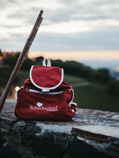 A red backpack is standing on a stone wall, next to a hiking pole. In the background, a beautiful evening sky can be seen, illuminating the city lights. | © Achromaticphotography