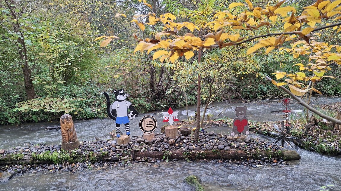 Eine kleine Bank am Ufer eines fließenden Gewässers mit mehreren bunten Holzfiguren. Im Hintergrund sind Bäume mit herbstlichen Blättern zu sehen. | © Edi Kager