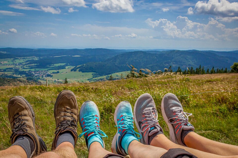 Eine Gruppe von Füßen in verschiedenen Schuhen sitzt auf einer Wiese. Im Hintergrund ist eine schöne Landschaft mit Hügeln und einem blauen Himmel zu sehen. | © pixabay.com