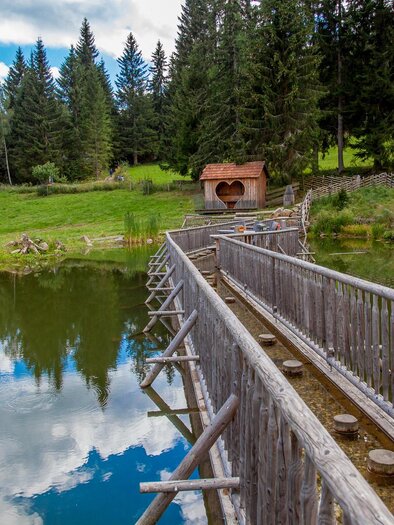 Eine Holzbrücke führt über einen ruhigen See, umgeben von hohen Bäumen. Im Hintergrund ist ein kleines Häuschen zu sehen, das die idyllische Landschaft ergänzt. | © Verein Romantischer Bründlweg