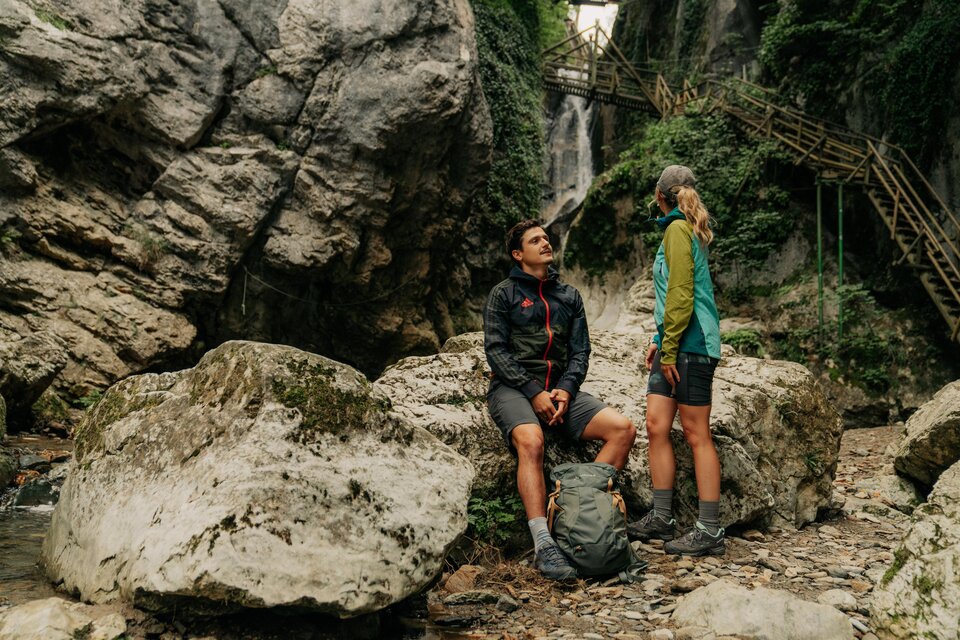 Zwei Wanderer machen eine Pause zwischen Felsen in der Kesselfallklamm, im Hintergrund Steg und Wasserfall. | © Region Graz-studio draussen