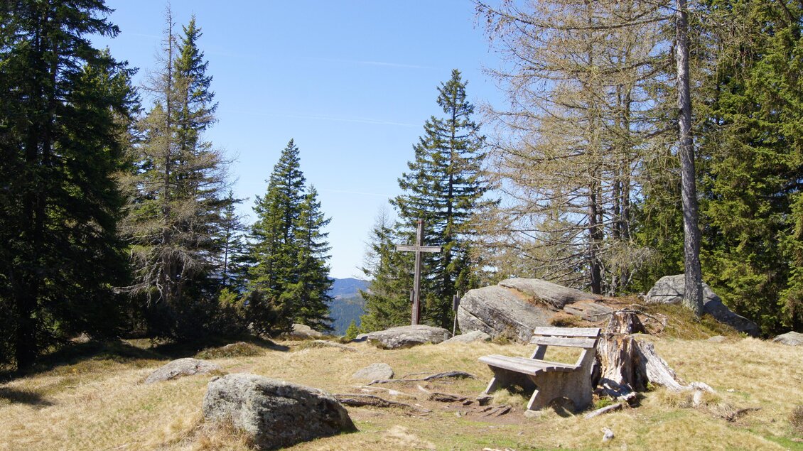 Gipfelkreuz am Reinischkogel mit Bank, Felsen und Nadelwald, Aussichtspunkt in der weststeirischen Berglandschaft. | © andikohlbacher