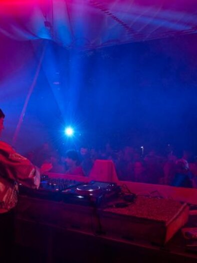 A DJ plays at a mixer in front of an excited crowd. Colorful lights and fog create a vibrant atmosphere. | © Monique_Fessl