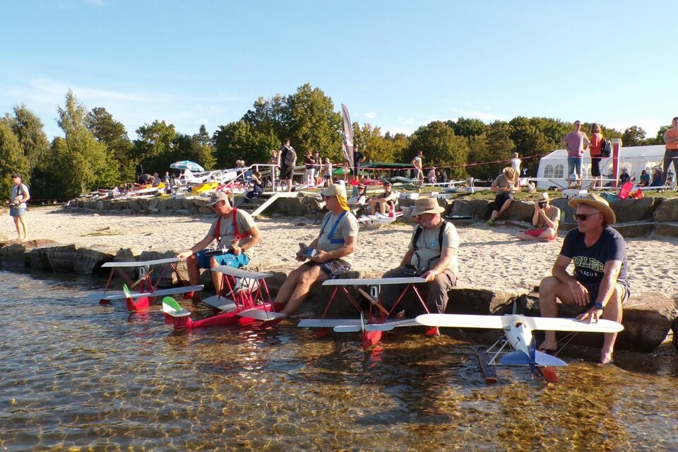 Gruppe von Männern, die an einem Strand mit ferngesteuerten Flugzeugen sitzen. Im Hintergrund sind mehrere Menschen und Bäume zu sehen. | © Modell Wasserflugverein Lausitz e. V.