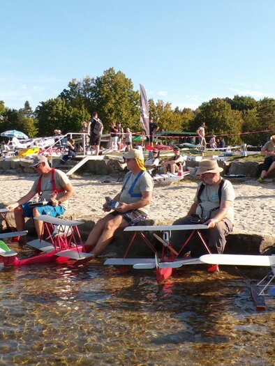 Group of men sitting on a beach with remote-controlled airplanes. In the background, several people and trees can be seen. | © Modell Wasserflugverein Lausitz e. V.