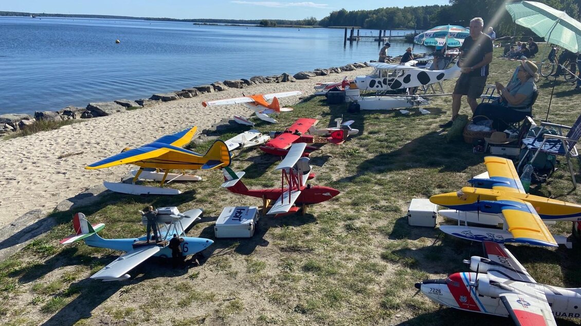Eine Reihe von Modellflugzeugen liegt am Ufer eines ruhigen Gewässers. Im Hintergrund sind Menschen zu sehen, die die Szene genießen. | © Modell Wasserflugverein Lausitz e. V.