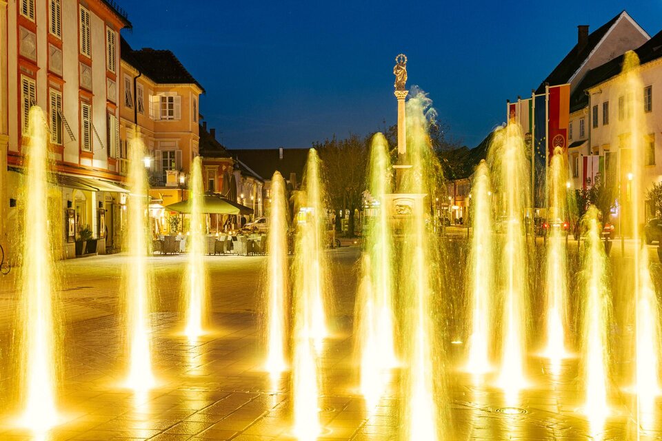Ein beleuchteter Brunnen auf einem Platz bei Nacht. Im Hintergrund sind Häuser und eine entspannte Atmosphäre sichtbar. | © © Thermen- & Vulkanland
