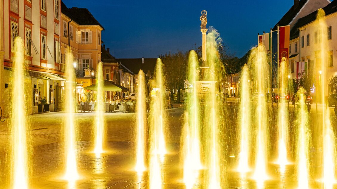 Ein beleuchteter Brunnen auf einem Platz bei Nacht. Im Hintergrund sind Häuser und eine entspannte Atmosphäre sichtbar. | © © Thermen- & Vulkanland