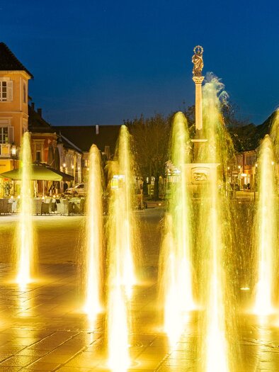 Ein beleuchteter Brunnen auf einem Platz bei Nacht. Im Hintergrund sind Häuser und eine entspannte Atmosphäre sichtbar. | © © Thermen- & Vulkanland