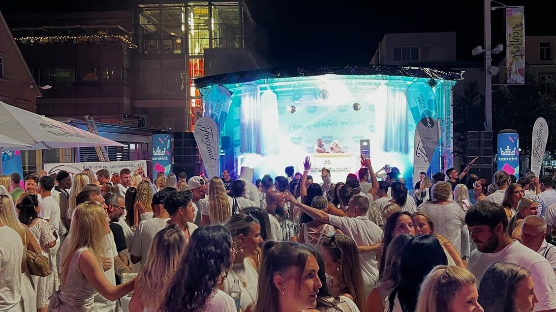 Eine lebhafte Nachtparty mit vielen Menschen in weißen Outfits. Im Hintergrund steht eine beleuchtete Bühne mit Musik und Unterhaltung. | © Segafredo Leoben