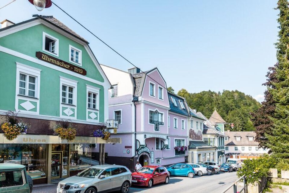 Eine malerische Straße mit bunten Häusern und Geschäften. Im Hintergrund sind grüne Hügel und ein blauer Himmel zu sehen.