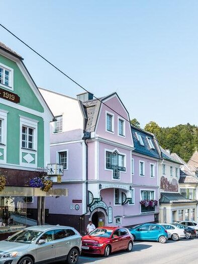 A picturesque street with colorful houses and shops. In the background, green hills and a blue sky can be seen.