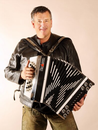 A man is holding an accordion and smiling warmly. He is wearing a leather jacket and has a relaxed look. | © Kirchenwirt Stubenberg