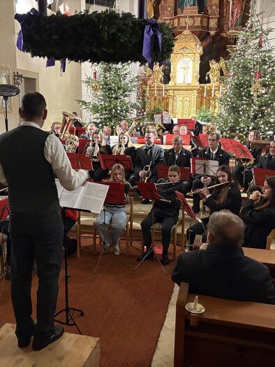 Weihnachtliche Klänge in festlicher Kirchenatmosphäre. Ein Blasorchester mit Musikerinnen und Musikern verschiedener Generationen musiziert unter der Leitung eines Dirigenten vor dem prachtvoll geschmückten Altar der Pfarrkirche Unterlamm. Beleuchtete Christbäume, ein großer Adventkranz und die stimmungsvolle Kulisse sorgen für eine besinnliche Atmosphäre und musikalische Vorfreude auf Weihnachten.

 | © MV Unterlamm