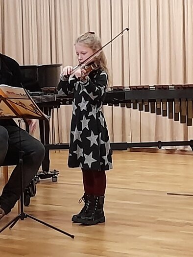 Ein junger Mann spielt Gitarre, während ein kleines Mädchen mit einer Violine musiziert. Im Hintergrund sind Musikinstrumente und eine Bühne zu sehen. | © Josef Fürstner