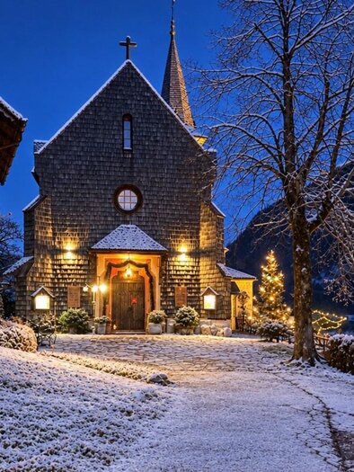 Eine charmante Kirche im Schnee, beleuchtet von warmem Licht. Die Umgebung ist malerisch mit schneebedeckten Bäumen und Bergen im Hintergrund. | © Bettina Scheck