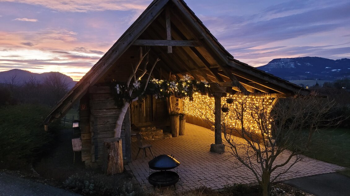 Eine charmante Holzhütte, beleuchtet von warmen Lichtern, vor einer malerischen Abenddämmerung. Im Hintergrund sind Berge und eine ruhige Landschaft zu sehen. | © Vital-Hotel-Styria