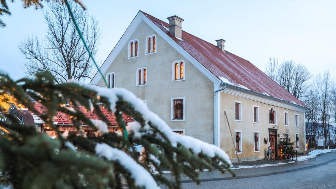 Ein charmantes Haus mit rotem Dach und weißen Wänden, umgeben von schneebedeckten Bäumen. Die Fenster des Hauses strahlen Wärme aus, während die kalte Winterlandschaft im Hintergrund sichtbar ist. | © © Foto De Monte