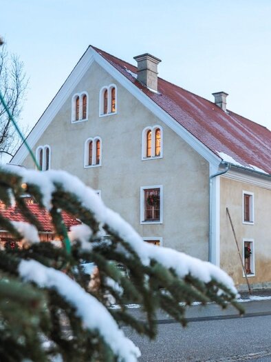 Ein charmantes Haus mit rotem Dach und weißen Wänden, umgeben von schneebedeckten Bäumen. Die Fenster des Hauses strahlen Wärme aus, während die kalte Winterlandschaft im Hintergrund sichtbar ist. | © © Foto De Monte