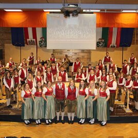 Eine große musizierende Gruppe in traditionellen österreichischen Trachten steht auf der Bühne. Im Hintergrund hängen Fahnen und ein Banner. | © Musikverein St. Lambrecht