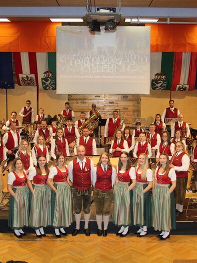 A large musical group in traditional Austrian costumes stands on the stage. In the background, flags and a banner are hanging. | © Musikverein St. Lambrecht