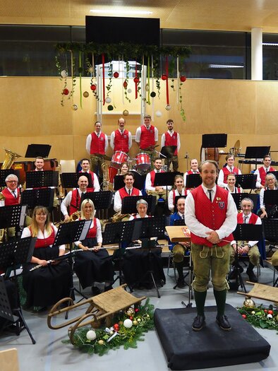 Eine große Blasmusikgruppe in traditionellen Trachten steht auf einer Bühne. Im Hintergrund hängen Weihnachtsdekorationen, und es sind Notenstände sichtbar. | © MV Weinitzen