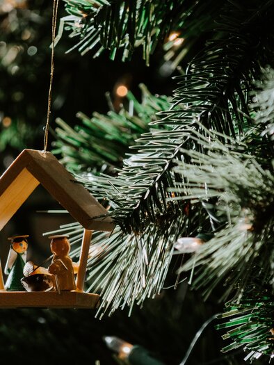 Ein Holzornament in Form eines Hauses hängt an einem Weihnachtsbaum. Darin stehen zwei Figuren, umgeben von grünen Ästen und Lichtern. | © TV Murau