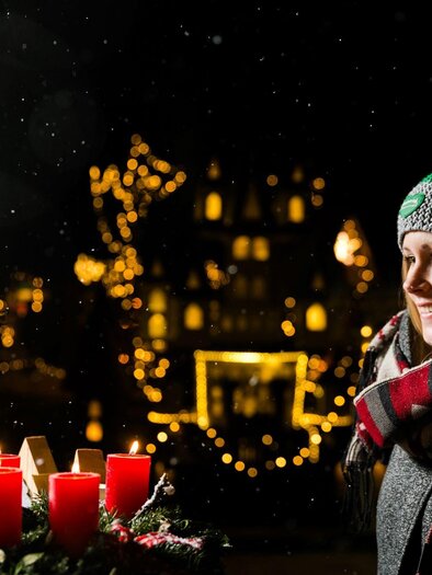 Eine Frau steht draußen und genießt die Weihnachtsstimmung bei Schneefall. Im Vordergrund brennen rote Kerzen und im Hintergrund leuchten festliche Lichter. | © TV Südsteiermark - Lupi Spuma