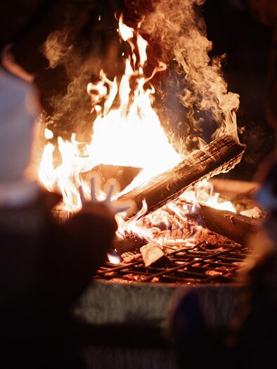 Ein gemütliches Lagerfeuer mit Flammen und glühenden Holzscheiten. Zwei Kinder sitzen in der Nähe und genießen die Wärme. | © Achromatic Photography