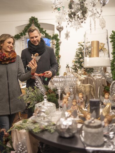 Ein gemütlicher Laden mit festlicher Weihnachtsdekoration. Ein Paar schaut sich Geschenke und Dekorationen an. | © Stadtgemeinde Feldbach I Bernhard Bergmann