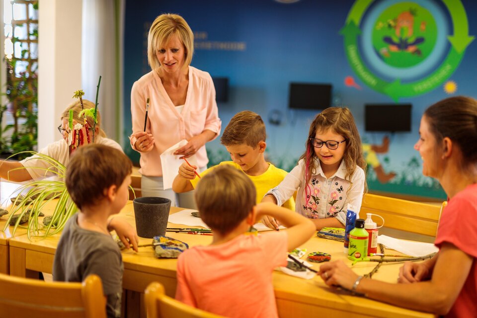 Eine Gruppe von Kindern und Erwachsenen sitzt an einem Tisch und arbeitet an einem kreativen Projekt. Eine Lehrerin erklärt den Kindern etwas, während sie gespannt zuhören. | © TV Murau