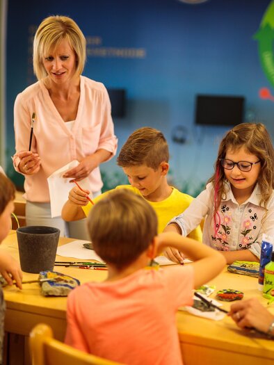 Eine Gruppe von Kindern und Erwachsenen sitzt an einem Tisch und arbeitet an einem kreativen Projekt. Eine Lehrerin erklärt den Kindern etwas, während sie gespannt zuhören. | © TV Murau