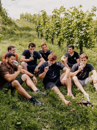 Eine Gruppe von sechs Männern sitzt auf dem Gras zwischen Weinreben. Sie genießen Getränke und unterhalten sich in einer entspannten Atmosphäre. | © sieme