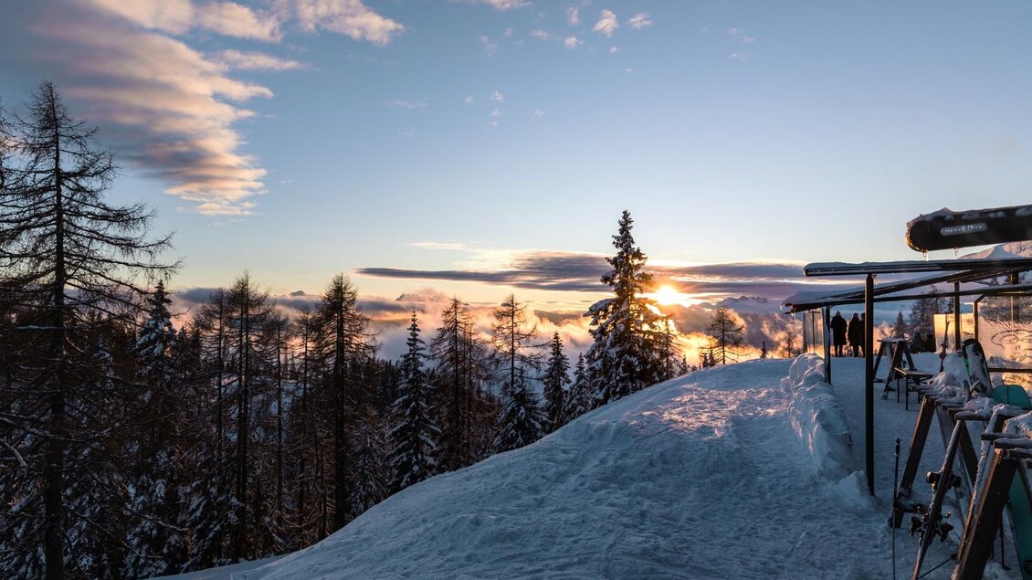 <p>Die winterliche Berglandschaft auf der Tauplitzalm mit schneebedeckten Bergen und Tannenbäumen. Die Sonne geht hinter den Bergen unter und taucht den Himmel in warme Farben. So mancher Skifahrer ist im Hollhaus eingekehrt und hat seine Ski vor dem Berggasthof stehen lassen.</p> | © needful.at und Berggasthof Hollhaus GmbH