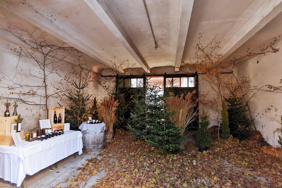 Ein rustikaler Raum mit Laub auf dem Boden und dekorierten Weihnachtsbäumen. Auf einem Tisch sind Getränke und Produkte ausgestellt. | © Tina Veit-Fuchs