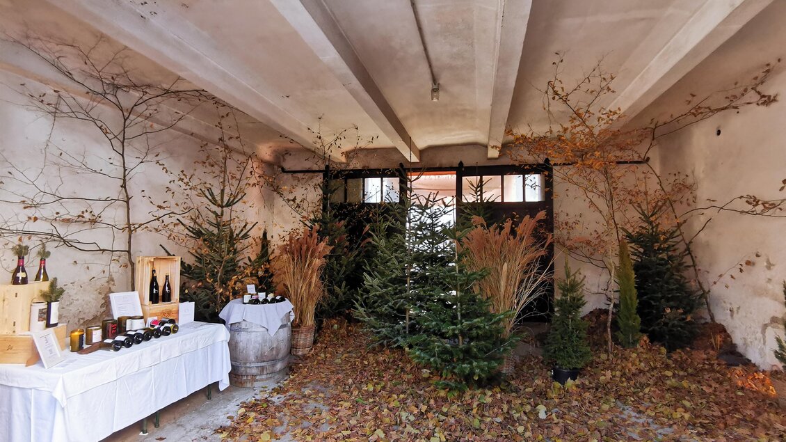 Ein rustikaler Raum mit Laub auf dem Boden und dekorierten Weihnachtsbäumen. Auf einem Tisch sind Getränke und Produkte ausgestellt. | © Tina Veit-Fuchs