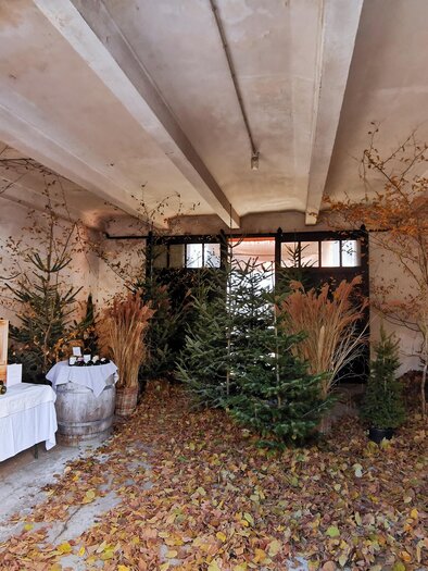 Ein rustikaler Raum mit Laub auf dem Boden und dekorierten Weihnachtsbäumen. Auf einem Tisch sind Getränke und Produkte ausgestellt. | © Tina Veit-Fuchs