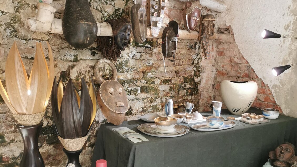 Eine schöne Ausstellung mit handgefertigten Kunstgegenständen. An der Wand hängen maskenartige Skulpturen, während auf einem Tisch verschiedene Dekorationsartikel präsentiert werden. | © Tina Veit-Fuchs