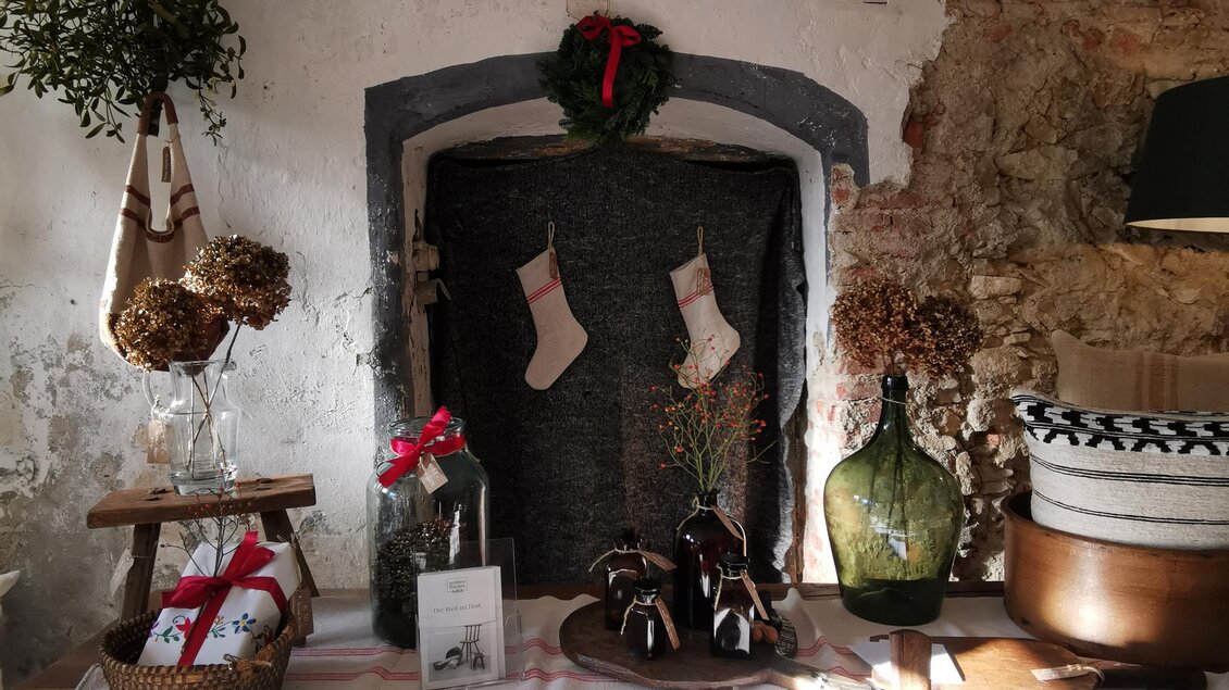 Ein gemütliches Zimmer mit rustikaler Einrichtung, in dem ein Tisch mit verschiedenen dekorativen Gegenständen steht. An der Wand hängen Weihnachtsstrümpfe und eine Kranzdekoration. | © Tina Veit-Fuchs