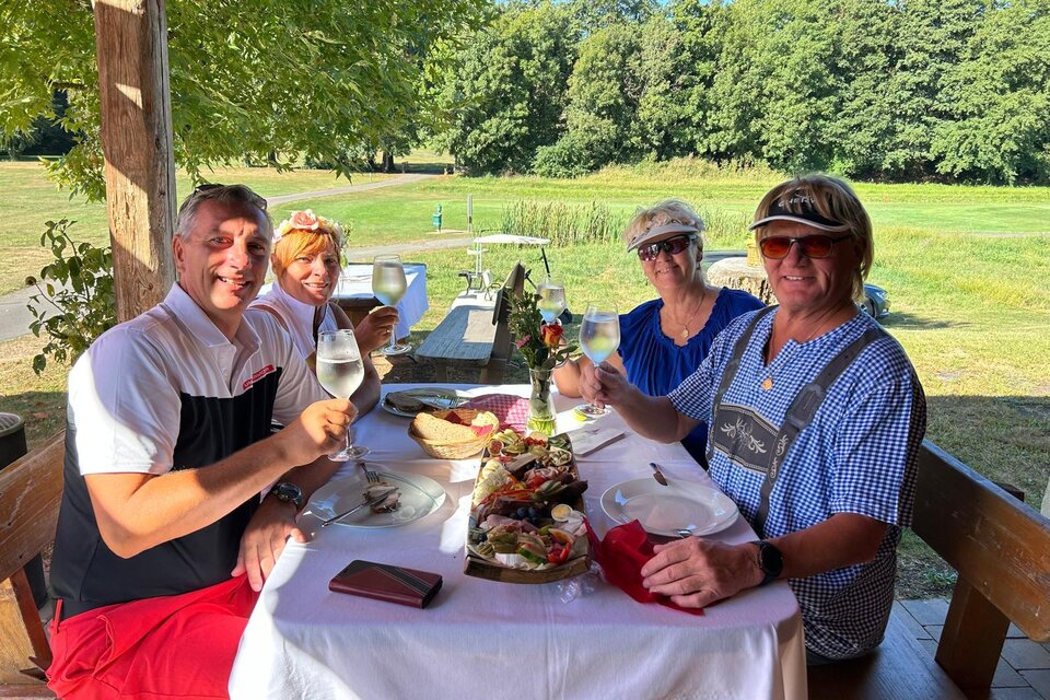Eine Gruppe von vier Personen sitzt an einem Tisch im Freien und hebt Gläser mit Getränken. Der Tisch ist schön gedeckt mit Essen im Hintergrund einer grünen Landschaft. | © Golf Traminer Klöch