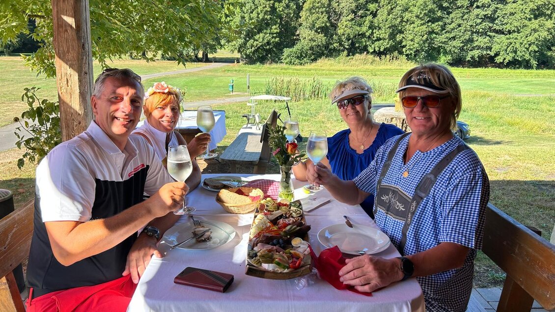 Eine Gruppe von vier Personen sitzt an einem Tisch im Freien und hebt Gläser mit Getränken. Der Tisch ist schön gedeckt mit Essen im Hintergrund einer grünen Landschaft. | © Golf Traminer Klöch