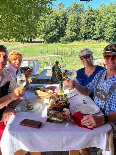 Eine Gruppe von vier Personen sitzt an einem Tisch im Freien und hebt Gläser mit Getränken. Der Tisch ist schön gedeckt mit Essen im Hintergrund einer grünen Landschaft. | © Golf Traminer Klöch