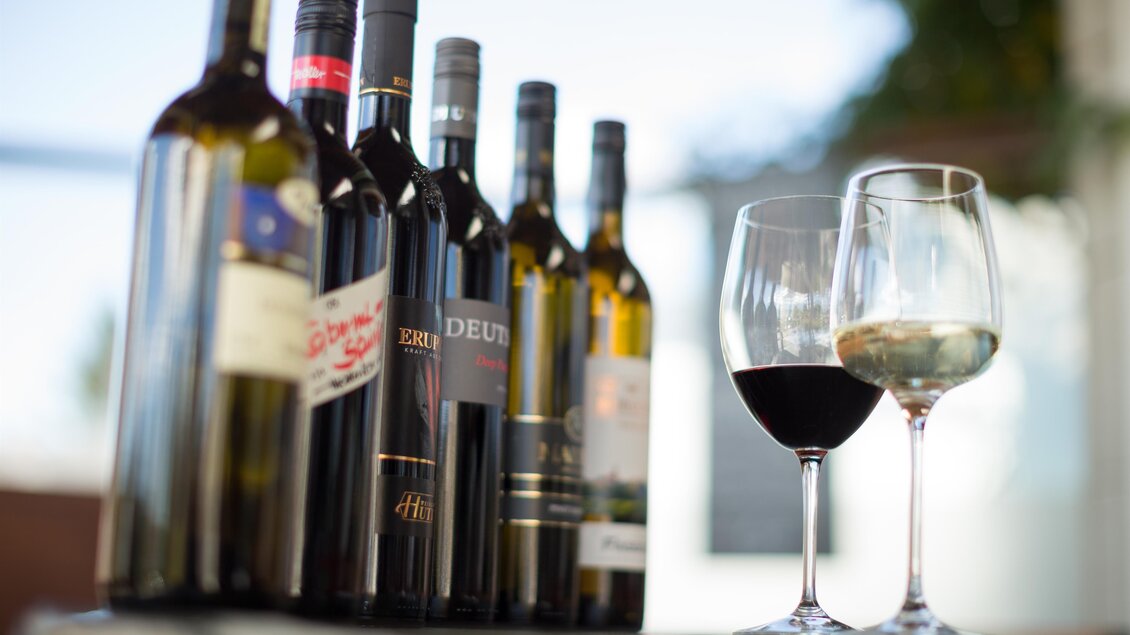 Eine Auswahl an Weinflaschen steht auf einem Tisch. Neben den Flaschen stehen zwei Weingläser, eines mit Rotwein und eines mit Weißwein.