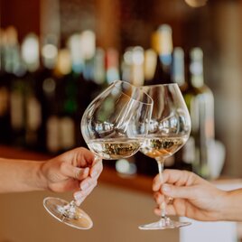 Two people are toasting with large wine glasses. In the background, many bottles of wine can be seen. | © @Karin Bergmann
