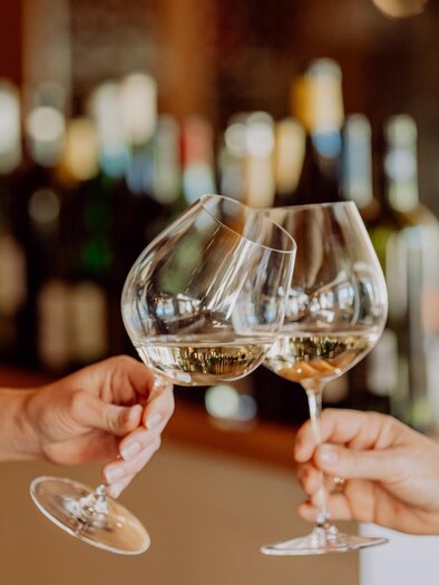 Two people are toasting with large wine glasses. In the background, many bottles of wine can be seen. | © Ratscher Landhaus