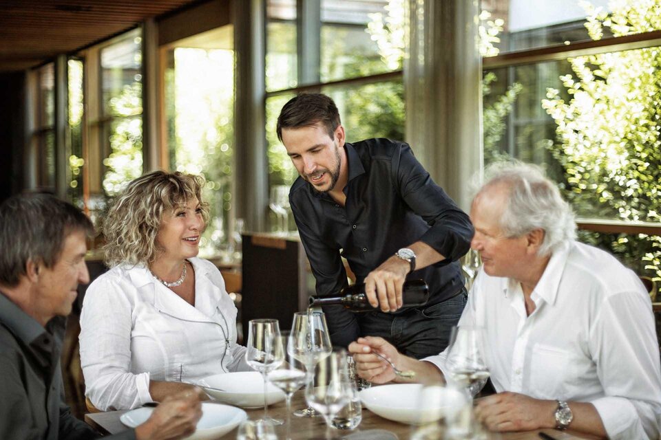 Eine Gruppe von vier Personen sitzt an einem Tisch in einem Restaurant. Ein Kellner serviert Wein, während die Gäste ansprechend miteinander kommunizieren. | © SIGNUM BLANC communications